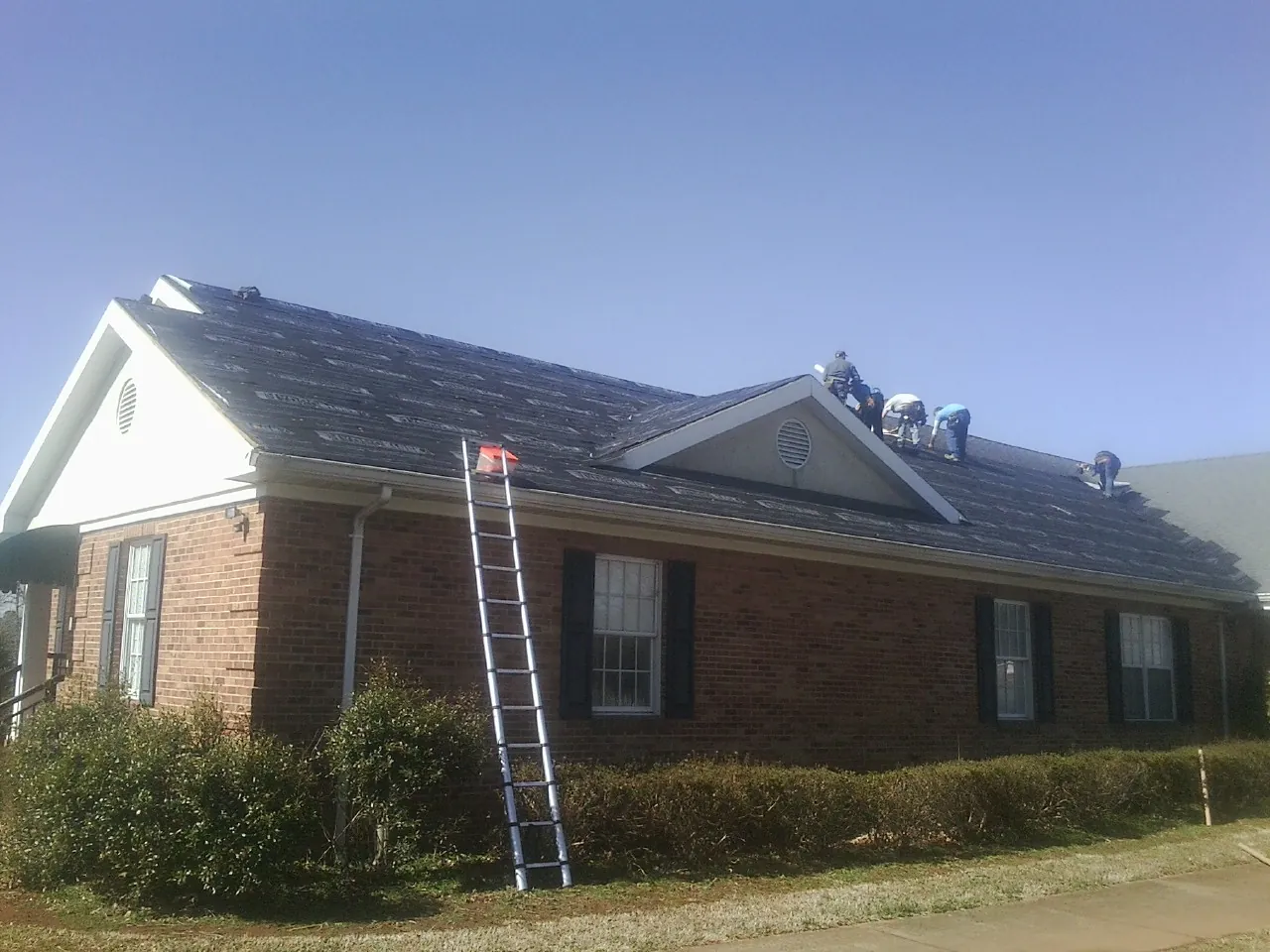 Crew tearing shingles off a brick ranch roof with a ladder set — Metal Roof Repair work by our crew in Bedias