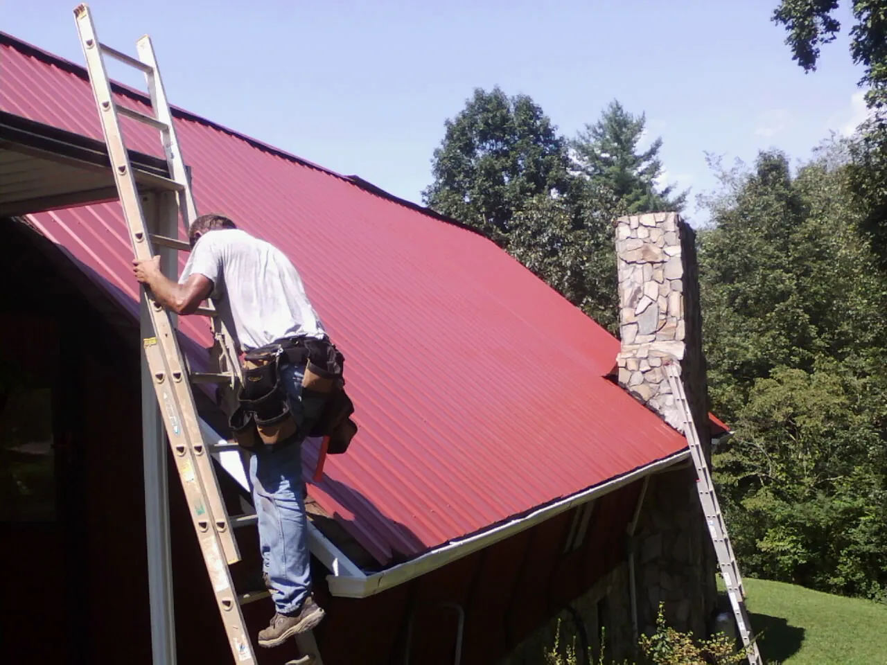 Roofer on a ladder beside a red metal roof and stone chimney — Soffit & Fascia Repair by Roofing Brothers in Bedias